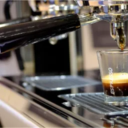 Kawa espresso spływająca do szklanki z profesjonalnego ekspresu ciśnieniowego z chromowanymi elementami, widoczna nazwa 'Espresso' na szklance.