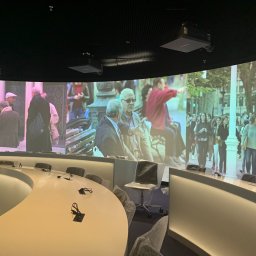 AB-INSTAL SPÓŁKA Z OGRANICZONĄ ODPOWIEDZIALNOŚCIĄ - Nowoczesna sala konferencyjna z panoramicznym ekranem projekcyjnym prezentującym scenę miejską. Krzesła i stoły w minimalistycznym stylu, mikrofony na stołach. Widoczne projektory sufitowe.