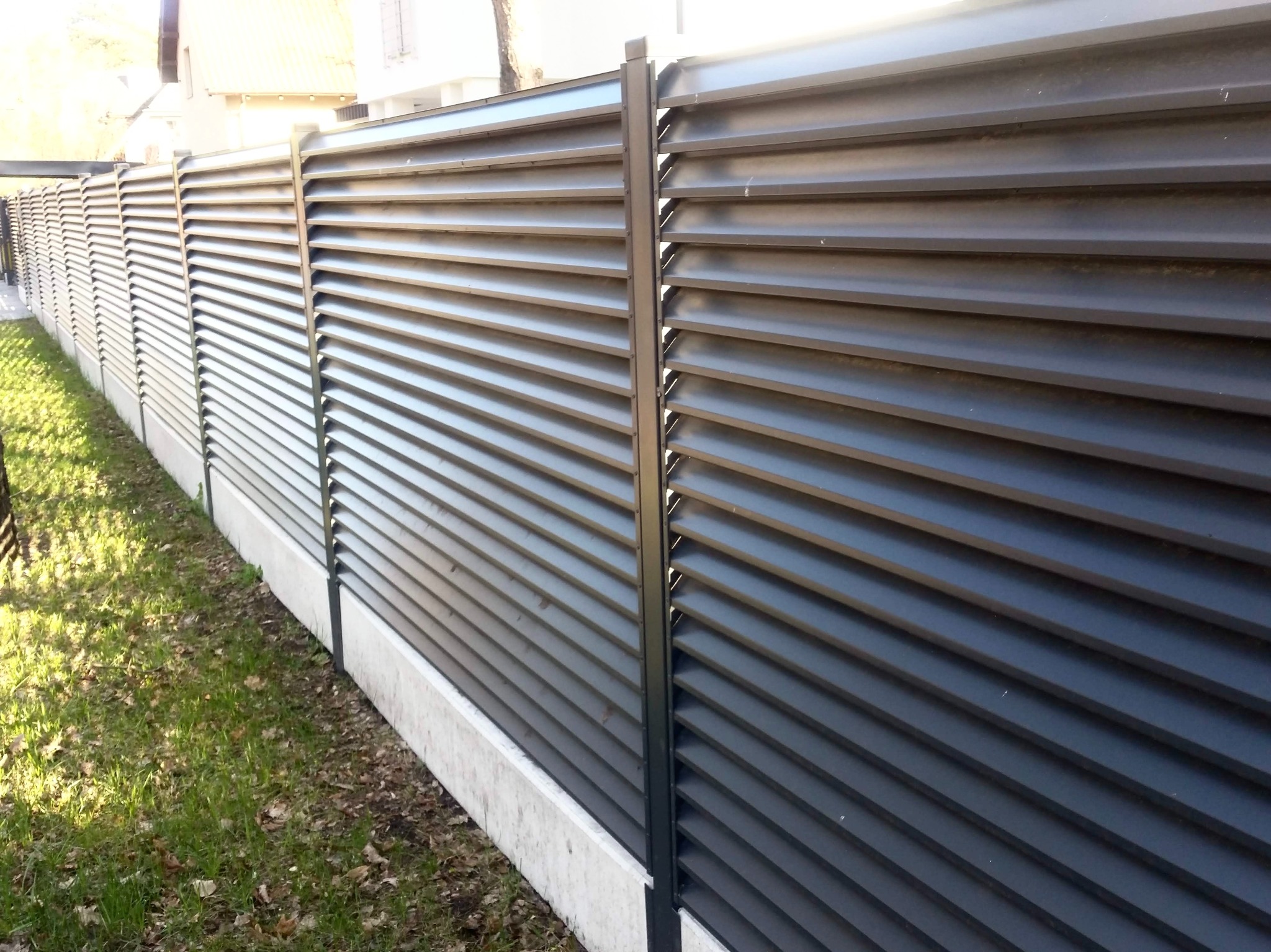 Nowoczesne, szare ogrodzenie lamelowe z betonową podmurówką, widziane z perspektywy bocznej, z fragmentem trawnika w tle. Minimalistyczny design.