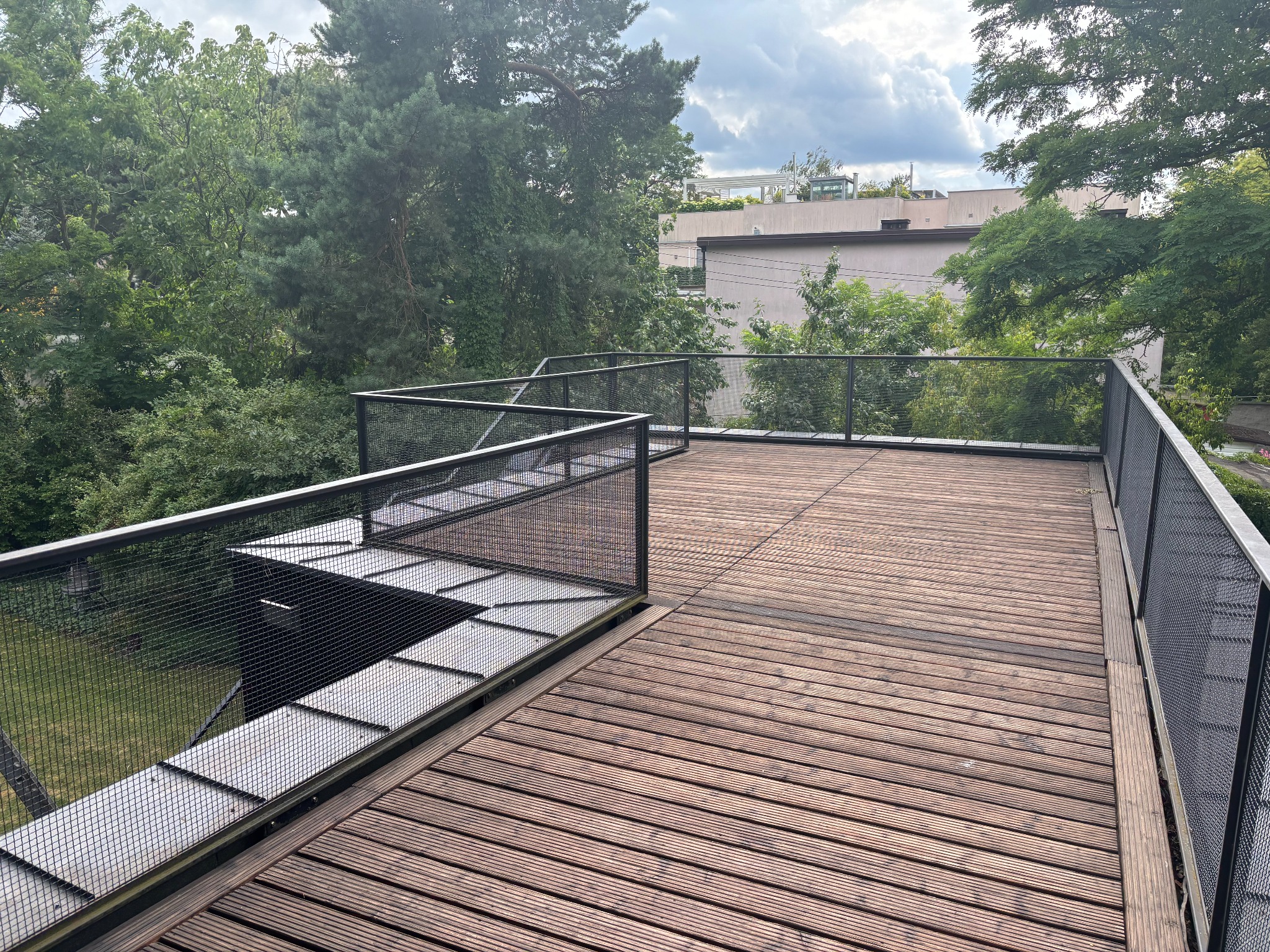 Drewniany taras z metalową balustradą z siatki na tle zieleni. Widok z góry, ukazujący strukturę desek i geometryczny wzór balustrady.