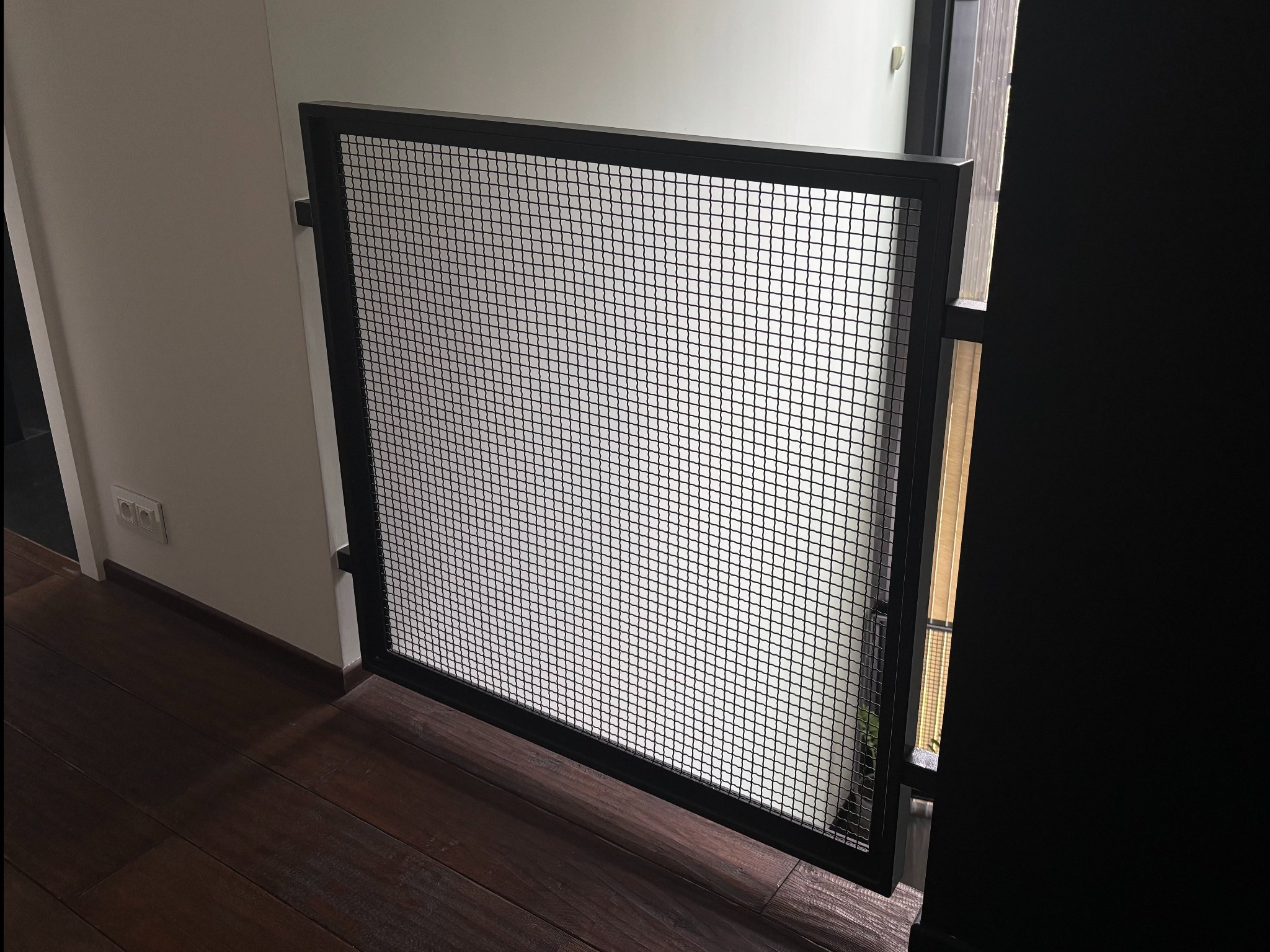 Czarna, metalowa barierka z siatki w nowoczesnym wnętrzu. Widoczne drewniane panele podłogowe i fragment ściany z gniazdkiem elektrycznym. Minimalistyczny design.