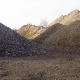 Góra żwiru, piasku i kamieni w słoneczny dzień. Widoczne ślady opon na ziemi. Różne frakcje kruszyw budowlanych składowane obok siebie.