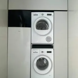 Kompaktowa instalacja: pompa ciepła Daikin obok pralki i suszarki w zabudowie meblowej, widoczny odpływ podłogowy i marmurowe płytki.