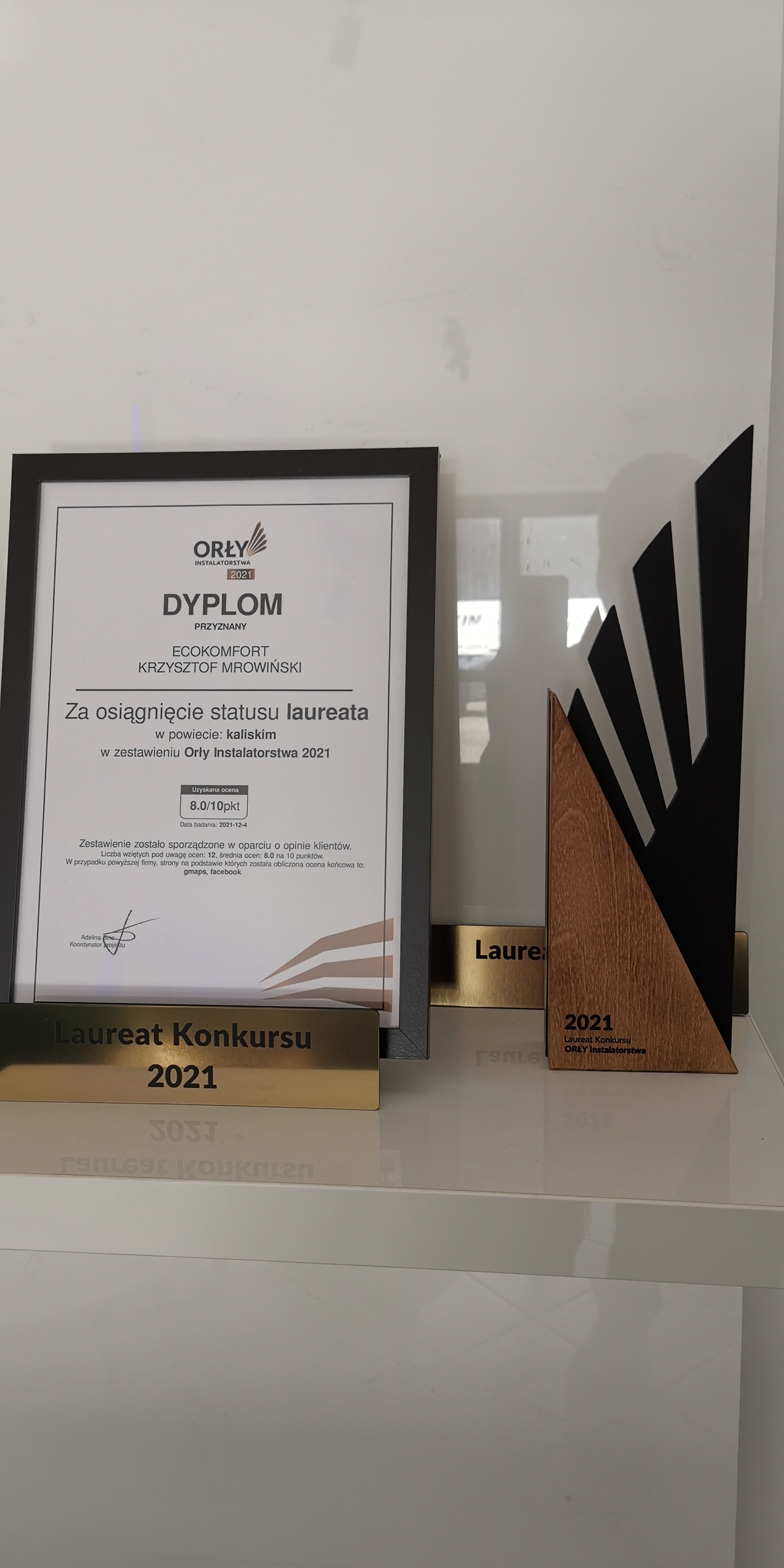 Dyplom i statuetka 'Laureat Konkursu Orły Instalatorstwa 2021' firmy EKO-KOMFORT Krzysztof Mrowiński, z oceną 8.0/10 pkt, na białym tle z odbiciami.