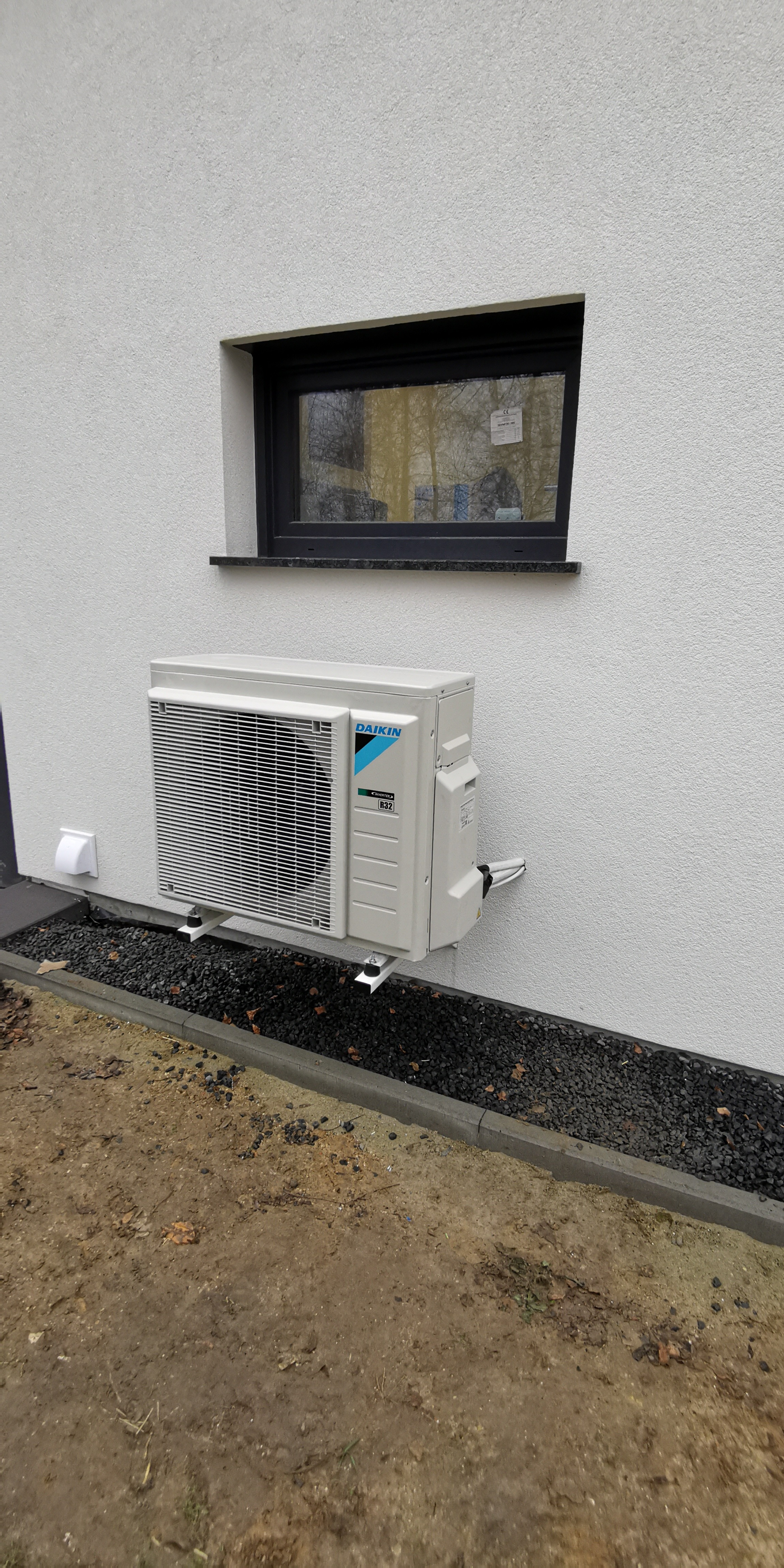 Zewnętrzna jednostka klimatyzacji Daikin zamontowana na białej ścianie budynku, widoczna mała czarna ramka okienna powyżej, czarne kamienie dekoracyjne u podstawy ściany.