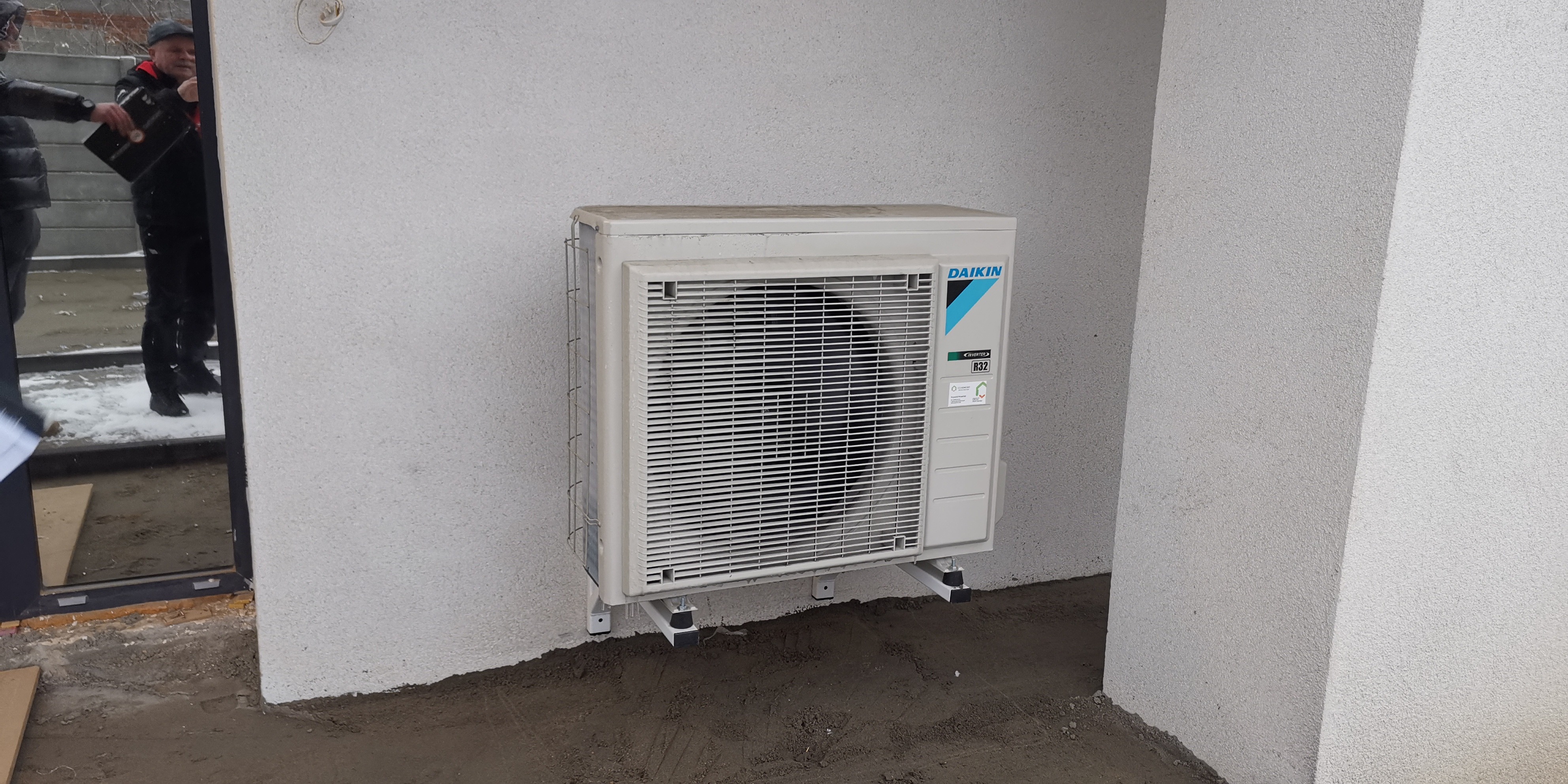 Zewnętrzna jednostka klimatyzacji Daikin zamontowana na szarej ścianie budynku z widocznym fragmentem okna i odbiciem postaci w szybie.