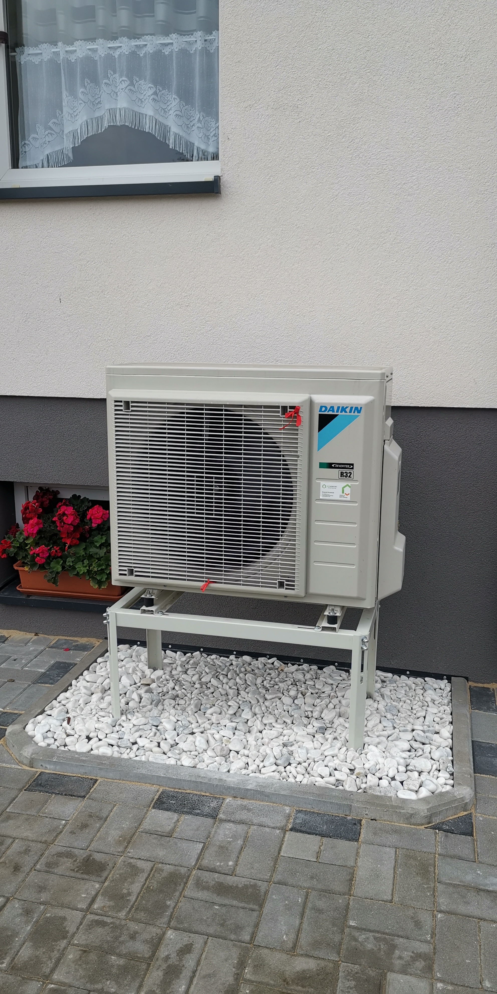 Zewnętrzna jednostka klimatyzacji Daikin R32 na metalowym stelażu, otoczona białymi kamieniami dekoracyjnymi, obok okna z firanką i donicą z czerwonymi kwiatami.