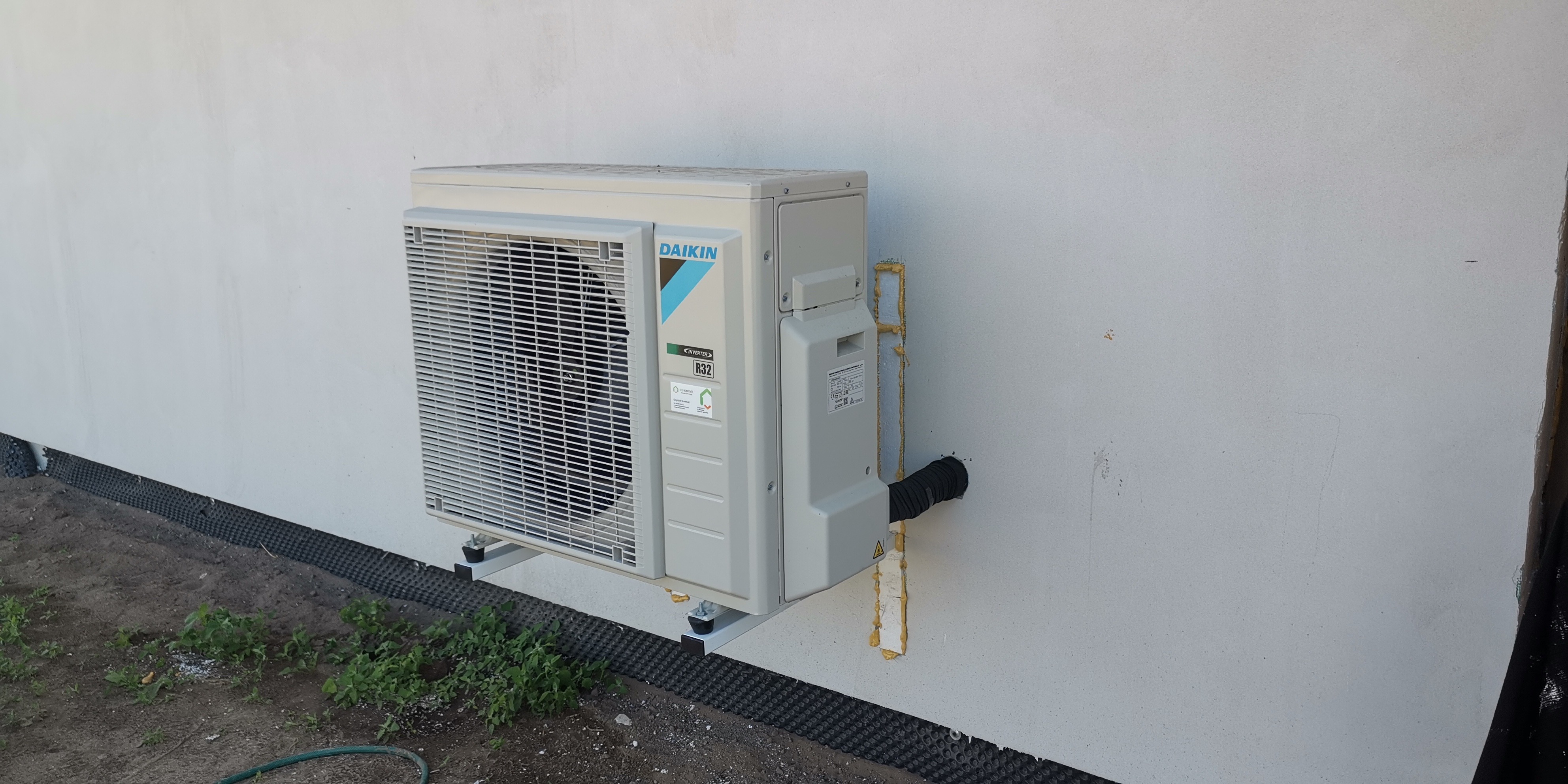 Zewnętrzna jednostka klimatyzacji Daikin zamontowana na ścianie budynku, widoczne rury instalacyjne zabezpieczone pianką montażową, pod jednostką fragment gruntu z roślinnością.
