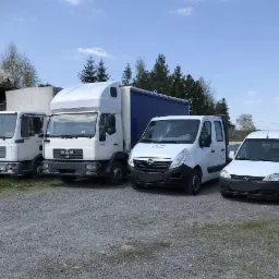 Flota transportowa: Dwa białe samochody ciężarowe MAN oraz dwa białe busy Opel na żwirowym placu, w tle budynek i drzewa pod błękitnym niebem.