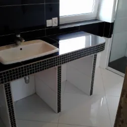 Nowoczesna łazienka z białą umywalką, czarnym blatem wykończonym mozaiką i czarnymi płytkami na ścianie. Minimalistyczny design z oknem w tle.