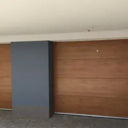 Dwie segmentowe bramy garażowe w kolorze jasnego drewna, zamontowane wewnątrz garażu z szarą kolumną i ścianami.