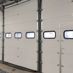 Zamknięte, segmentowe bramy przemysłowe w kolorze białym, z poziomymi przetłoczeniami i małymi okienkami w czarnych ramkach, widok z wnętrza hali.