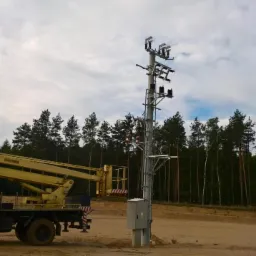 Żółty podnośnik koszowy przy betonowym słupie energetycznym z rozdzielnią, na tle lasu i pochmurnego nieba.