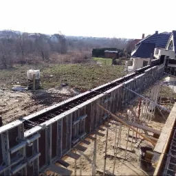 Szczegółowy widok szalunku zbrojeniowego pod wylewkę fundamentów domu jednorodzinnego na działce z widokiem na okoliczną zabudowę.