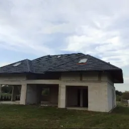 Surowy stan domu jednorodzinnego z szarą dachówką i oknami dachowymi, widoczny z zewnątrz na tle pochmurnego nieba.