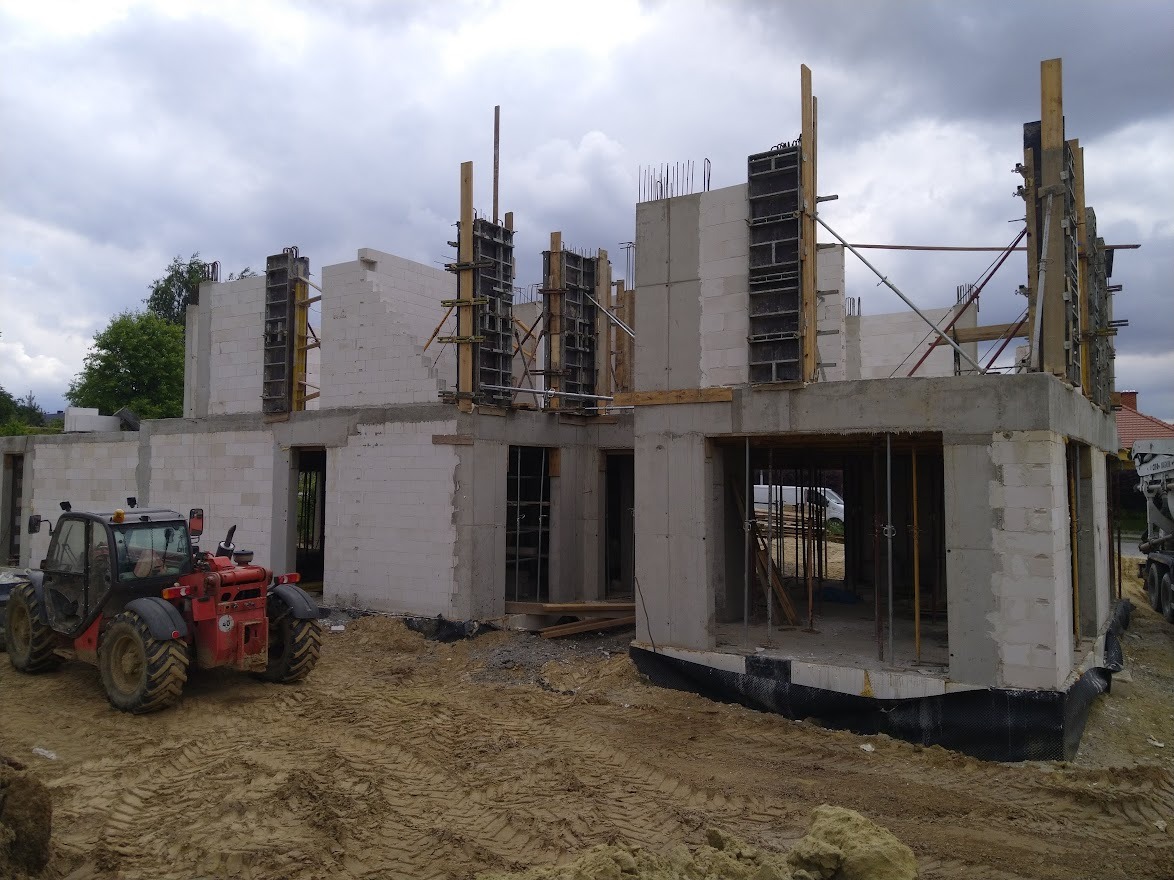 Budowa domu murowanego z betonu komórkowego, widoczny podnośnik teleskopowy i szalunki na ścianach, teren budowy z wyraźnymi śladami opon.