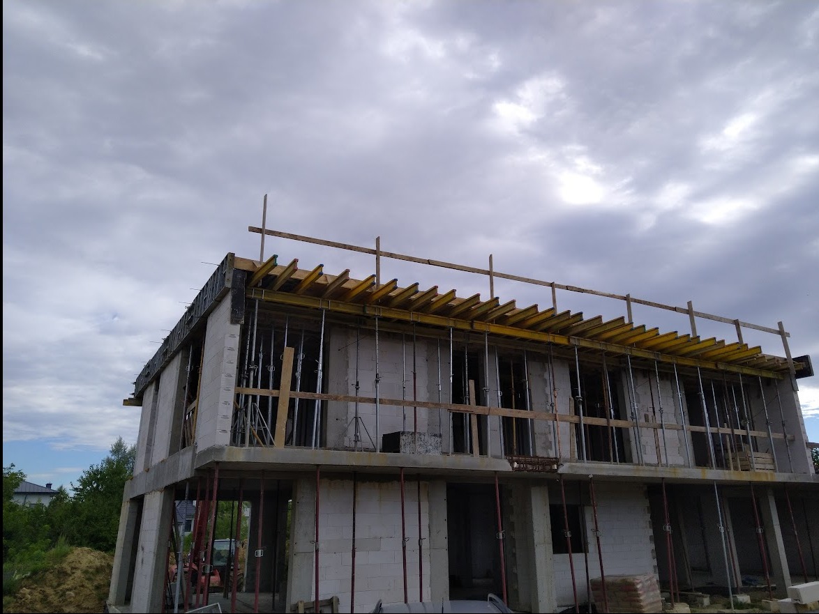 Surowy stan dwukondygnacyjnego domu z betonu komórkowego w trakcie budowy, widoczne podpory szalunkowe, drewniane belki stropowe, zbrojenia i szary, zachmurzony krajobraz w tle.