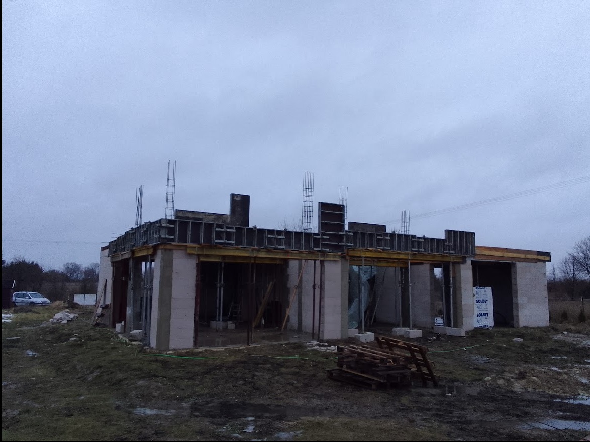 Budowa domu jednorodzinnego w stanie surowym otwartym; widoczne ściany z bloczków, szalunki stropowe zbrojone prętami oraz składowane palety na zewnątrz.