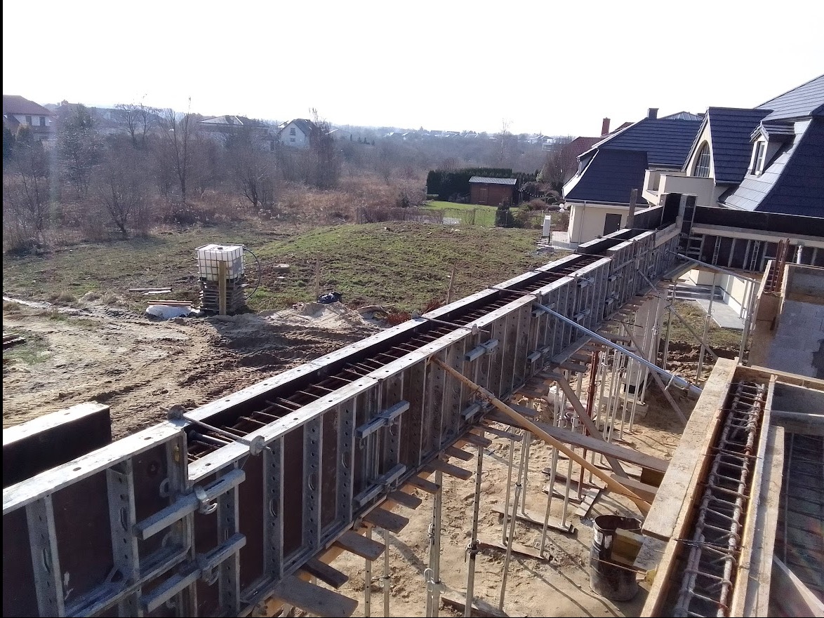 Szczegółowy widok szalunku zbrojeniowego pod wylewkę fundamentów domu jednorodzinnego na działce z widokiem na okoliczną zabudowę.