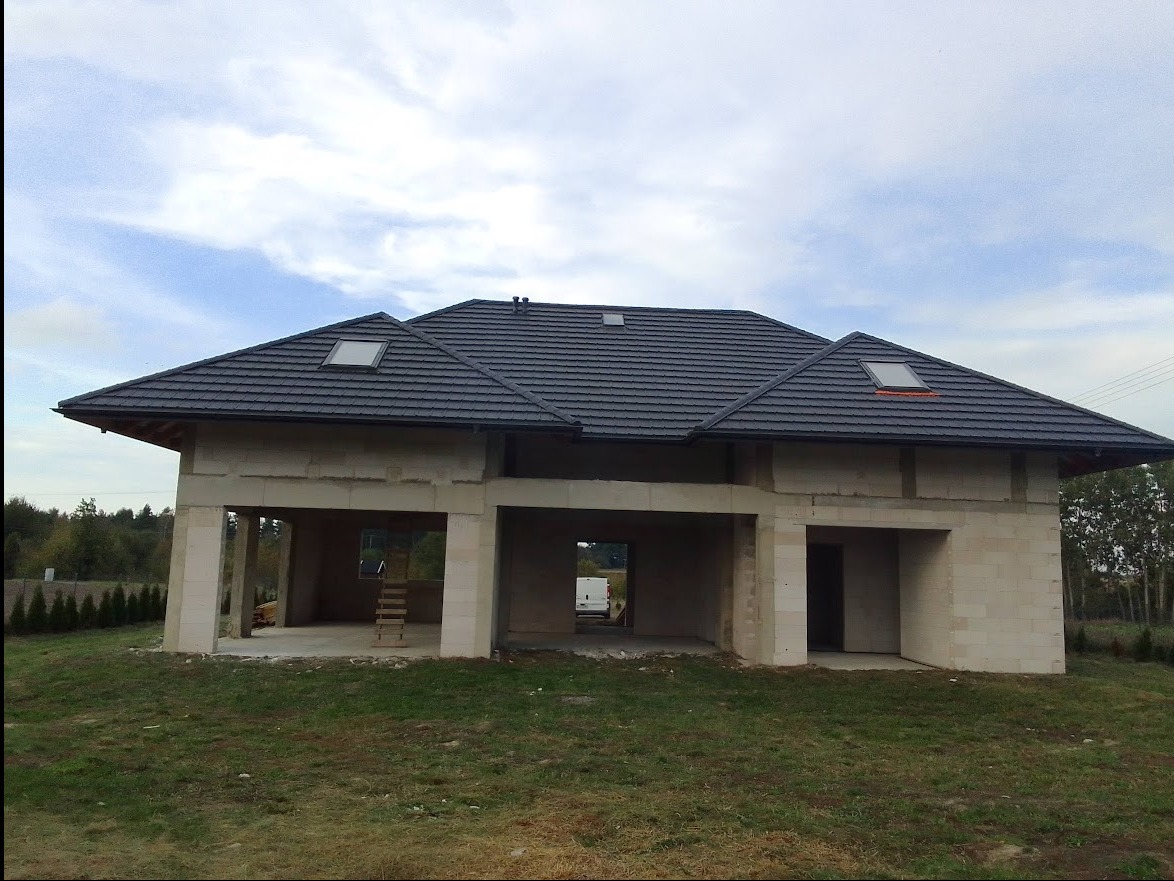 Surowy stan domu jednorodzinnego z szarej cegły, z ciemnym dachem z dachówką i oknami dachowymi, widok z zewnątrz na niedokończone wnętrze z drabiną.