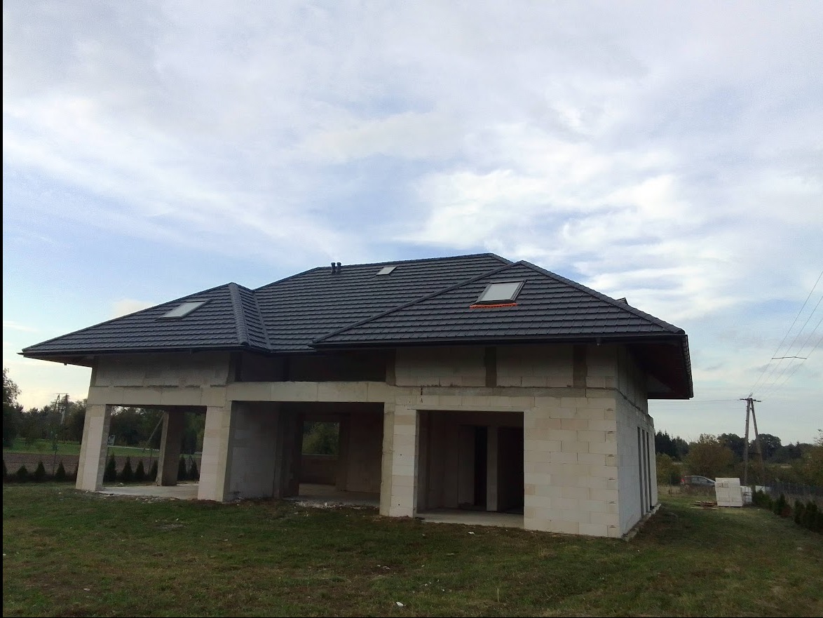 Surowy stan domu jednorodzinnego z szarą dachówką i oknami dachowymi, widoczny z zewnątrz na tle pochmurnego nieba.