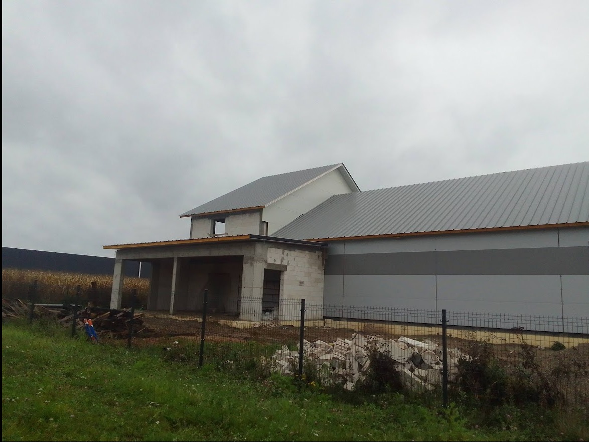 Surowy stan budowy domu z betonu komórkowego, widoczna konstrukcja dachu i ściany szczytowe, na pierwszym planie ogrodzenie i gruz budowlany, pochmurne niebo w tle.