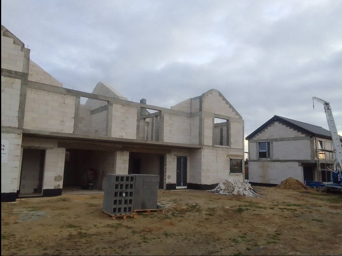 Dwa domy w stanie surowym otwartym, zbudowane z białych bloczków, na jednym z nich widoczna czarna hydroizolacja fundamentów, a w tle widoczny podnośnik koszowy.