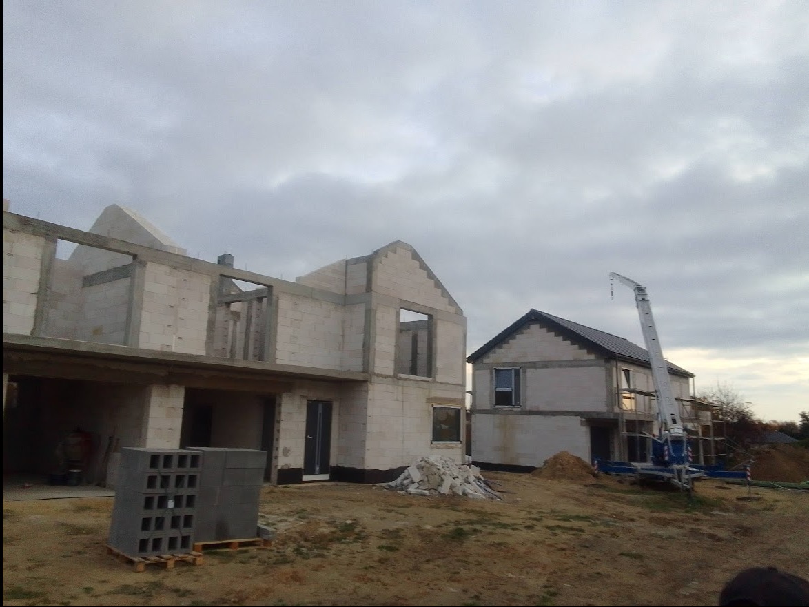 Budowa dwóch domów z białych pustaków, jeden w stanie surowym otwartym z widocznym zbrojeniem, drugi z dachem i oknami, na placu budowy stoi dźwig.