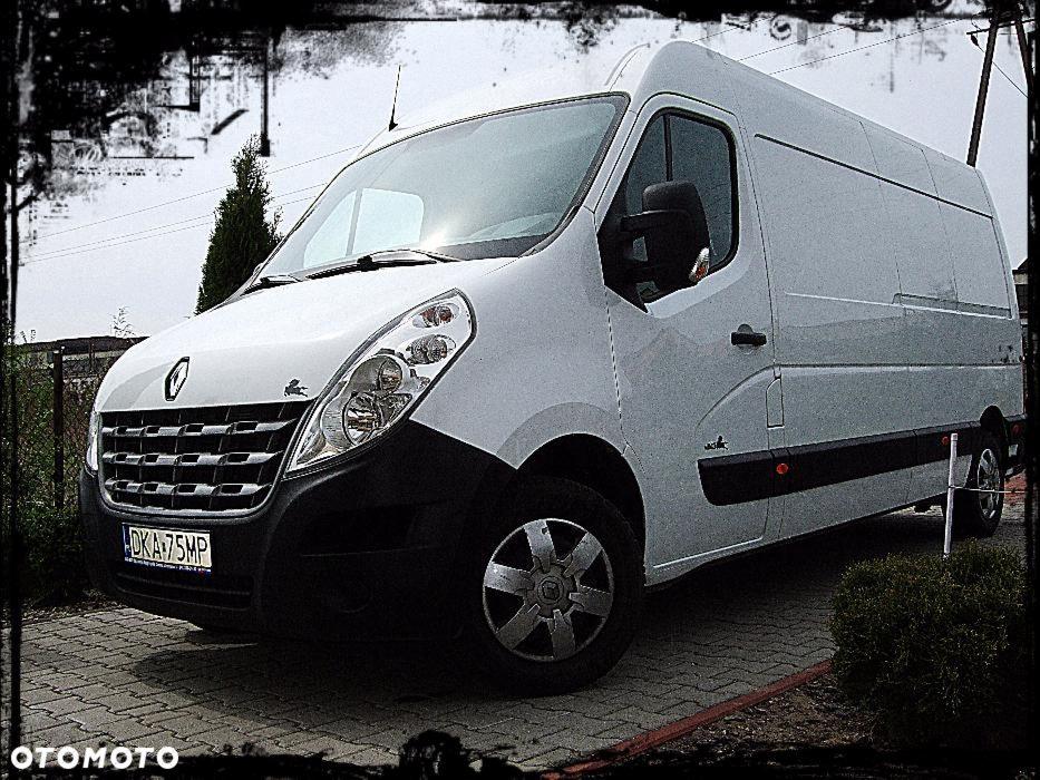 Renault Master w kolorze białym, zaparkowany na kostce brukowej. Widoczny z boku pod kątem, z numerem rejestracyjnym DKA 75MP. Tło stanowią krzewy i ogrodzenie.