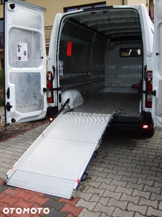 Wnętrze białego busa z otwartymi drzwiami i rozłożoną rampą ułatwiającą załadunek, widok od tyłu, na zewnątrz kostka brukowa.