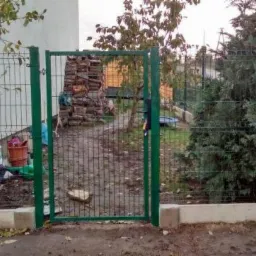 Otwarta zielona furtka w ogrodzeniu z siatki panelowej, widok na stos drewna i iglaki w tle, z lewej strony narzędzia ogrodnicze opierają się o ogrodzenie.