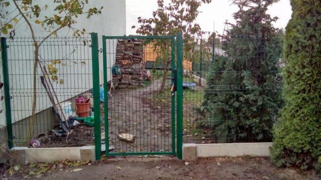 Otwarta zielona furtka w ogrodzeniu z siatki panelowej, widok na stos drewna i iglaki w tle, z lewej strony narzędzia ogrodnicze opierają się o ogrodzenie.