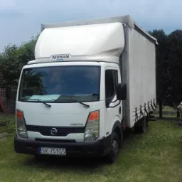 Biały samochód dostawczy Nissan Cabstar z plandeką, zaparkowany na trawie, z widoczną tablicą rejestracyjną 'SK 751GS'.