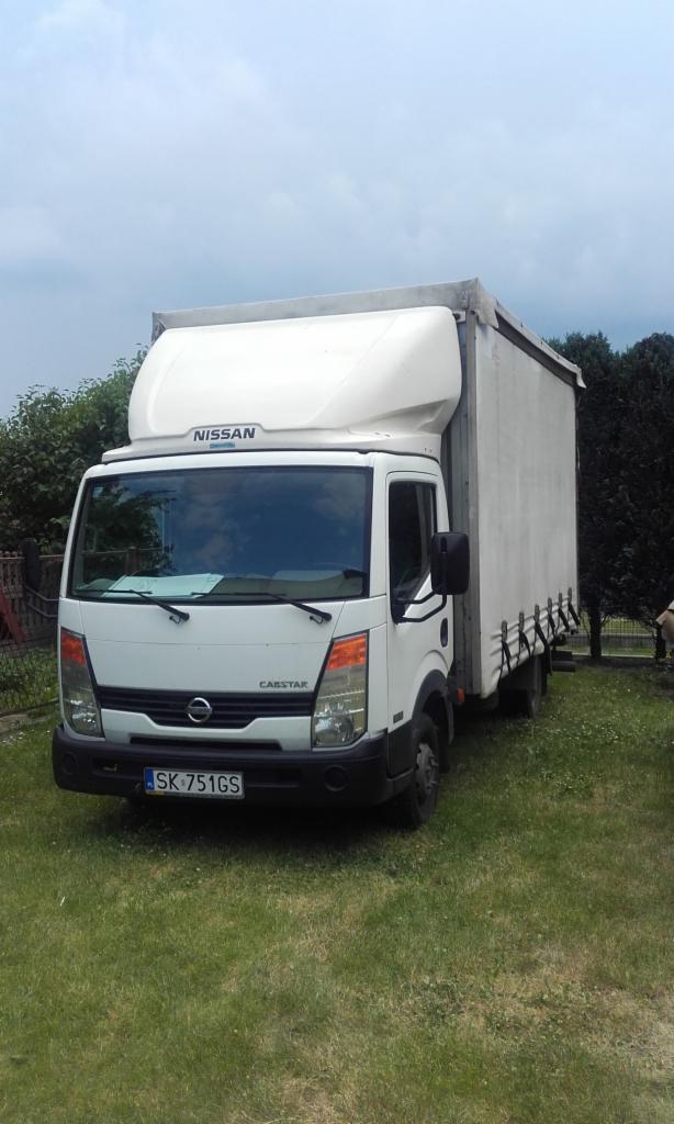 Biały samochód dostawczy Nissan Cabstar z plandeką, zaparkowany na trawie, z widoczną tablicą rejestracyjną 'SK 751GS'.