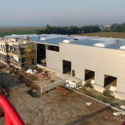 M.J.J MET Sp z o.o - Wysoki kąt widzenia na budowę hali przemysłowej z widocznymi oknami dachowymi i pracownikami na dachu, z fragmentem rusztowania i izolacji na sąsiednim budynku biurowym w tle krajobraz polny.