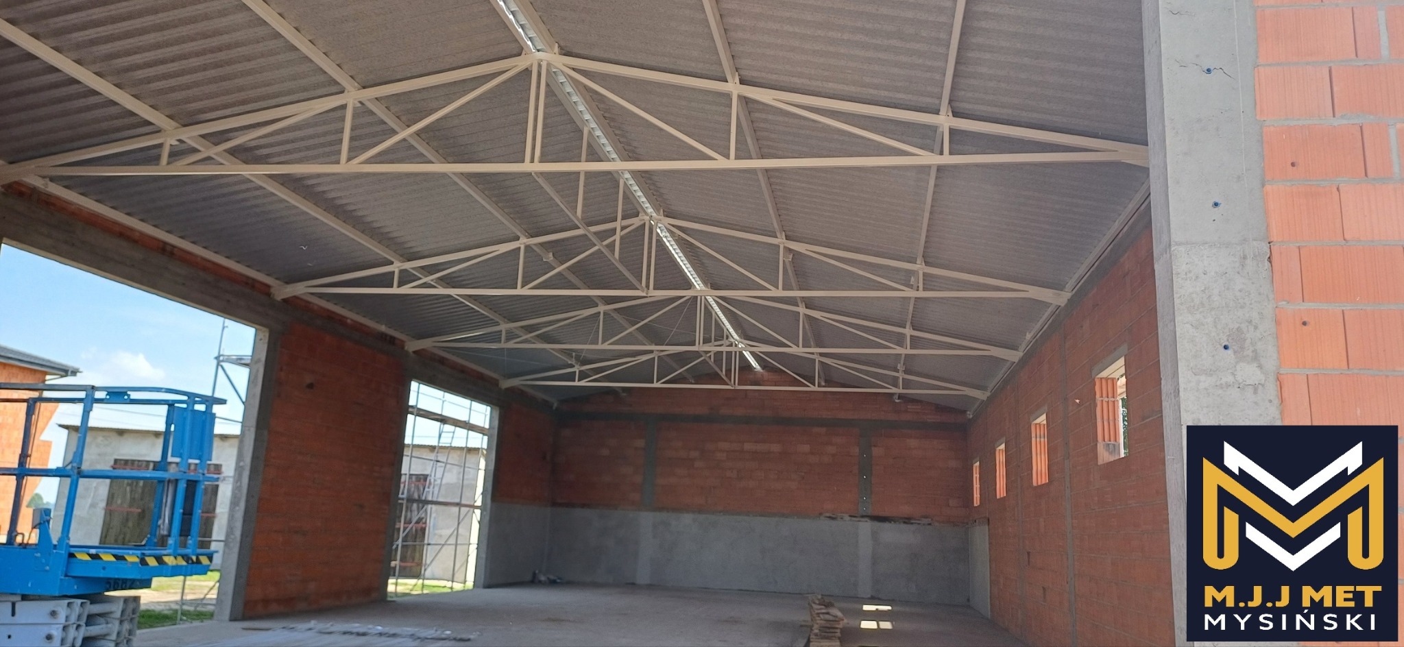 Wnętrze hali w trakcie budowy, widoczne stalowe elementy konstrukcyjne dachu, ściany z czerwonej cegły oraz podnośnik koszowy na zewnątrz.