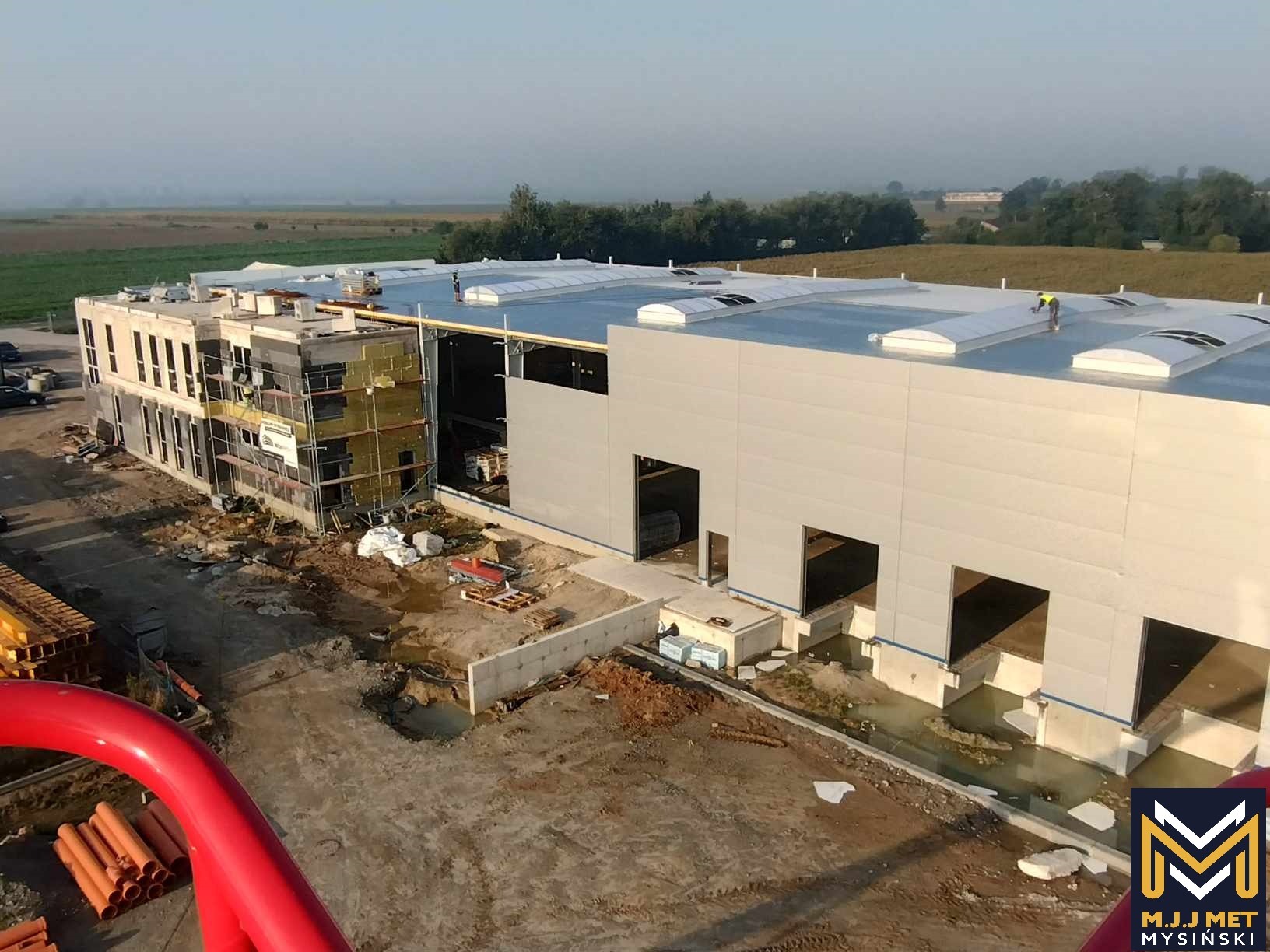 Wysoki kąt widzenia na budowę hali przemysłowej z widocznymi oknami dachowymi i pracownikami na dachu, z fragmentem rusztowania i izolacji na sąsiednim budynku biurowym w tle krajobraz polny.