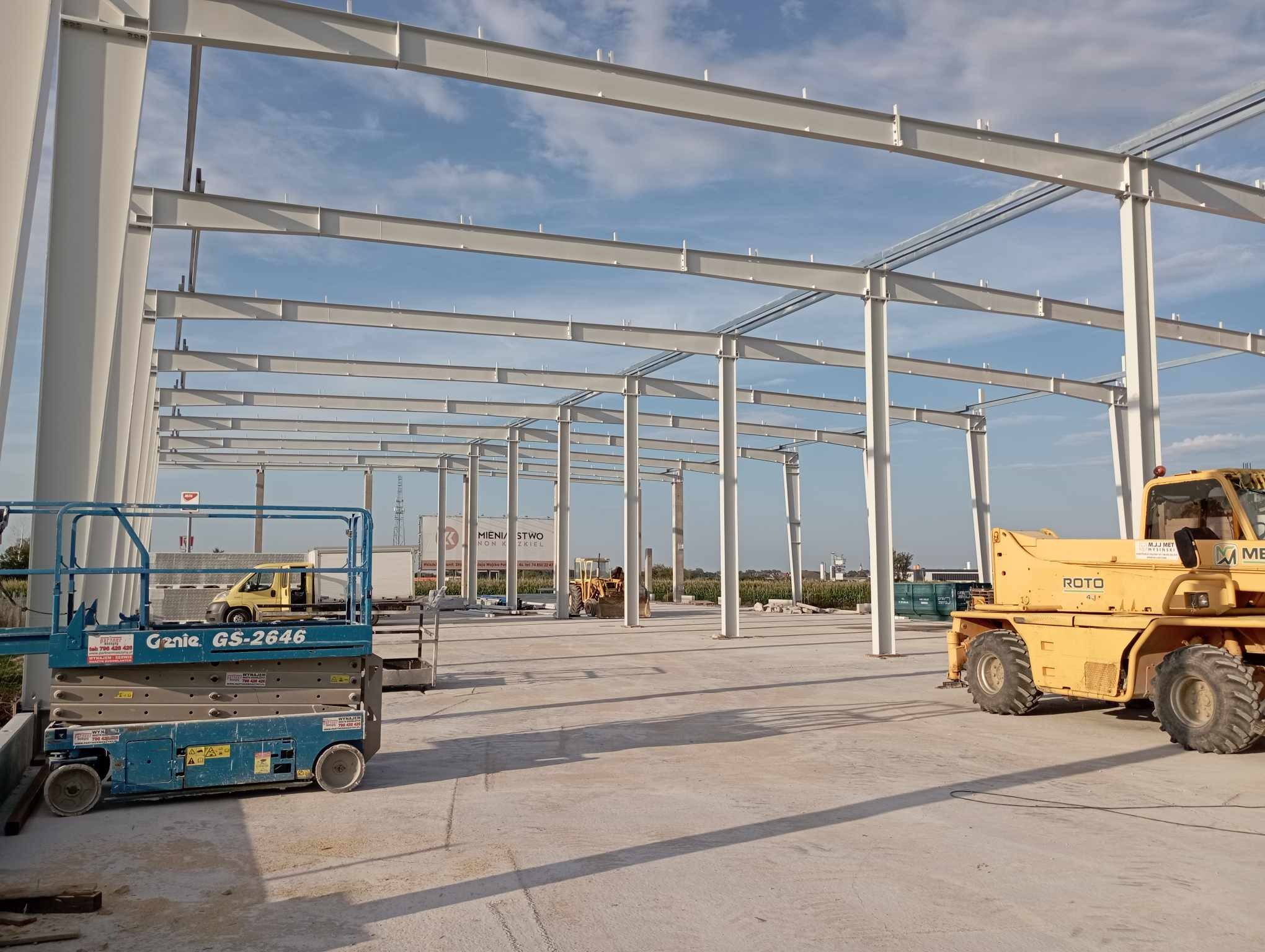 Konstrukcja hali w trakcie budowy, widoczne stalowe elementy nośne, podnośnik nożycowy Genie GS-2646 i żółta ładowarka teleskopowa ROTO 4.1 na betonowej posadzce, błękitne niebo w tle.