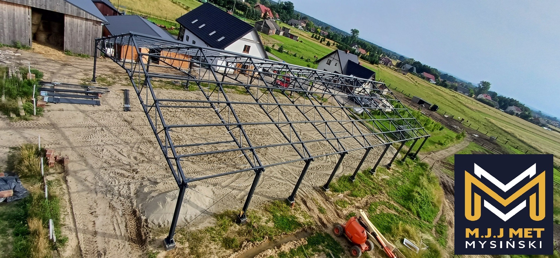 Szkielet konstrukcji stalowej hali w trakcie budowy na piaszczystym terenie, widok z góry na wieś i pola, z boku leżą przygotowane elementy konstrukcyjne.