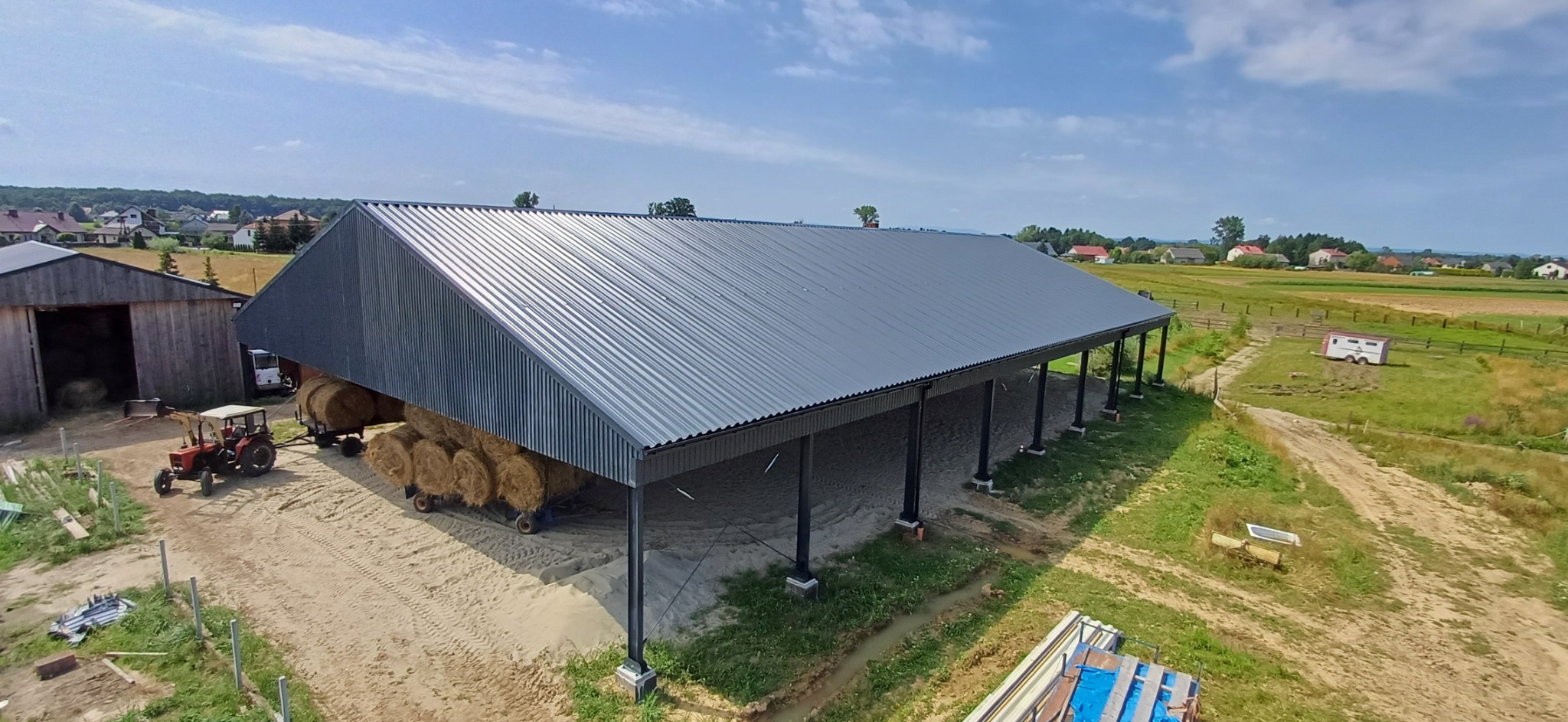 Zadaszona konstrukcja stalowa z szarej blachy trapezowej, wsparta na słupach, służąca jako wiata na siano, z traktorem i przyczepą w tle, na wiejskim krajobrazie.