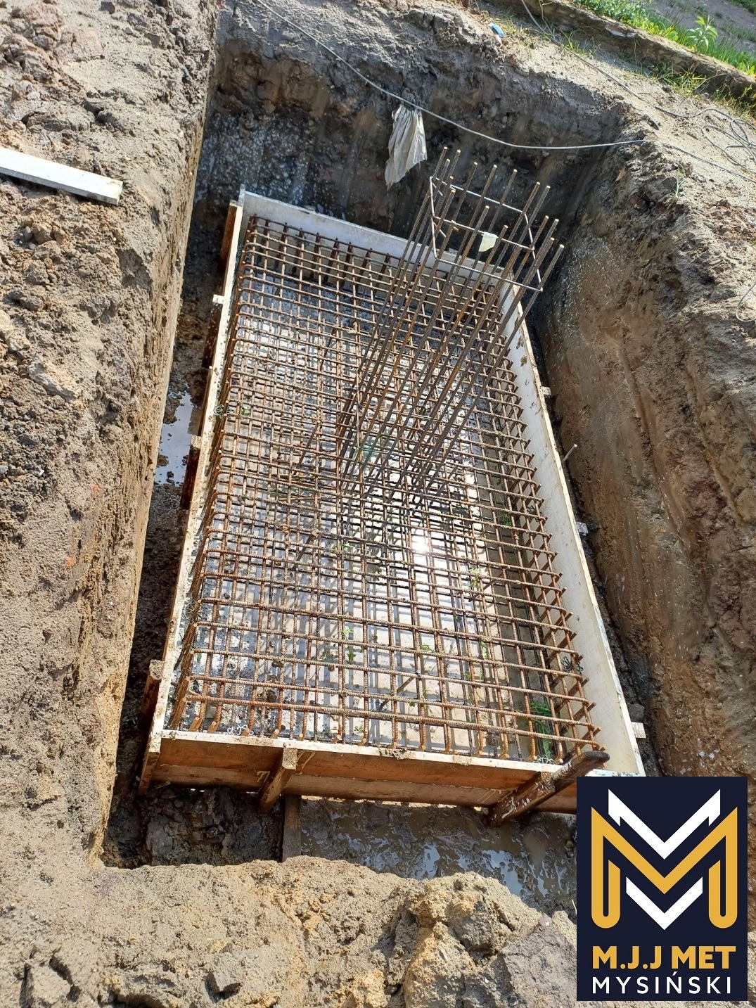 Zbrojenie fundamentu w wykopie, siatka stalowa z pionowymi prętami, szalunek drewniany, logo firmy w prawym dolnym rogu.