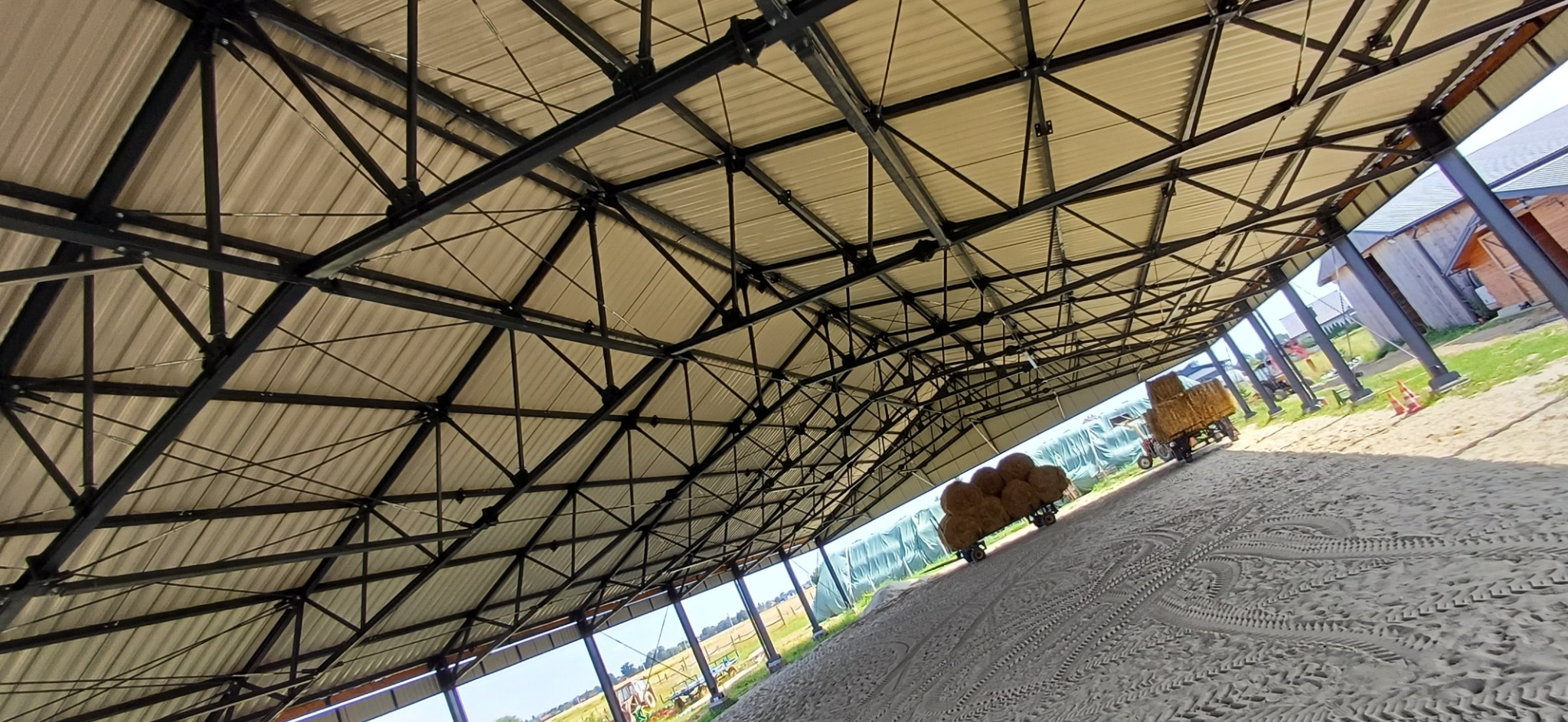 Wnętrze hali z widoczną konstrukcją stalową dachu, w tle wóz z belami siana na piaszczystym podłożu.