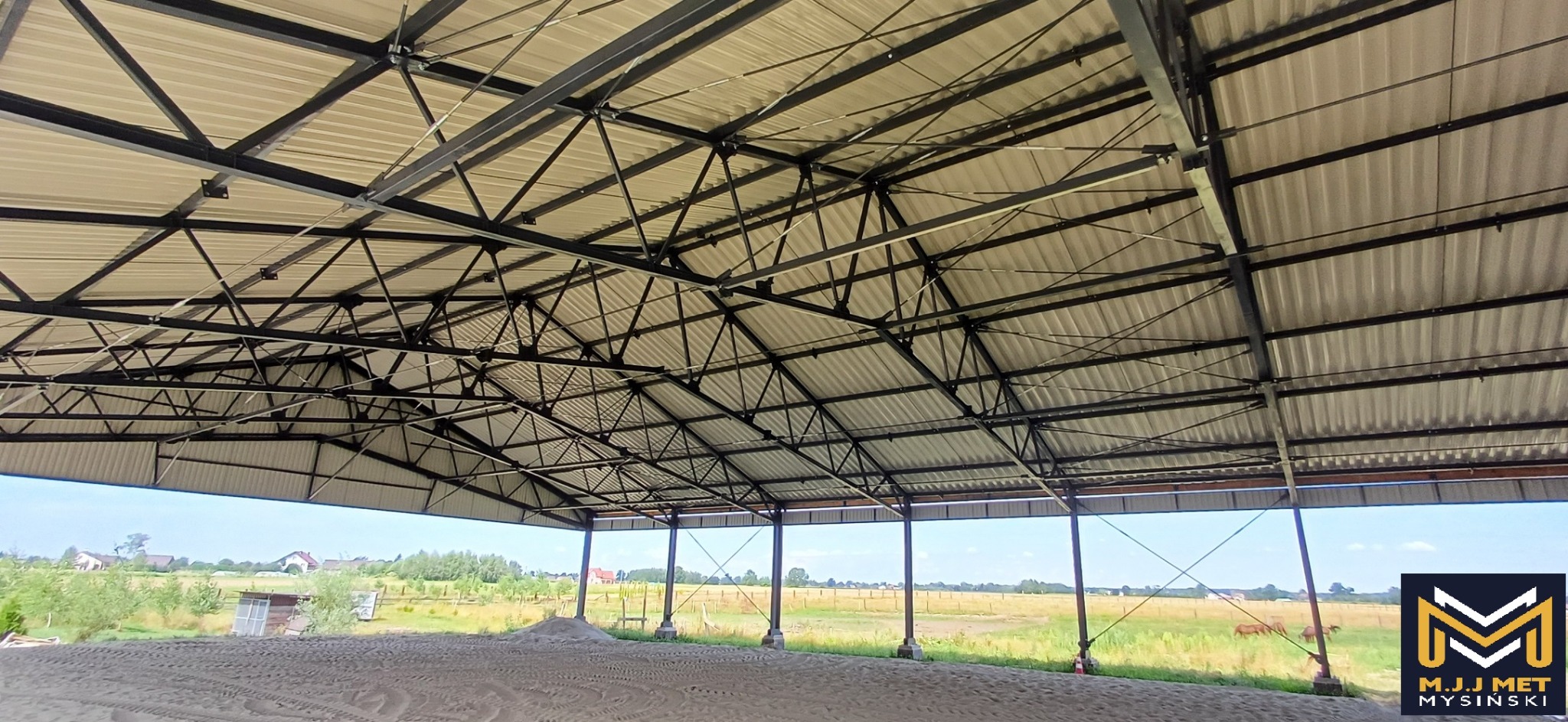Wnętrze hali z konstrukcją stalową dachu, widok na zewnątrz na łąkę i zabudowania, z logo firmy w prawym dolnym rogu.
