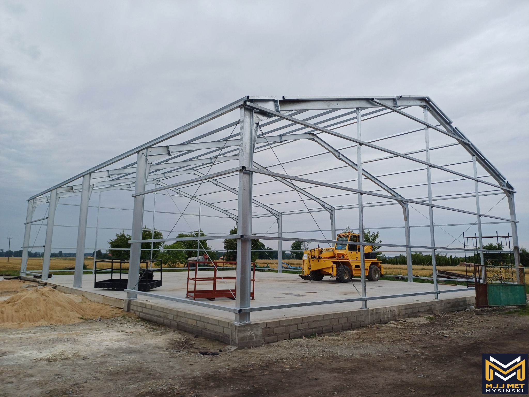 Szkielet powstającej hali stalowej na betonowym fundamencie, z żółtym wózkiem widłowym w tle i stertą piasku obok.