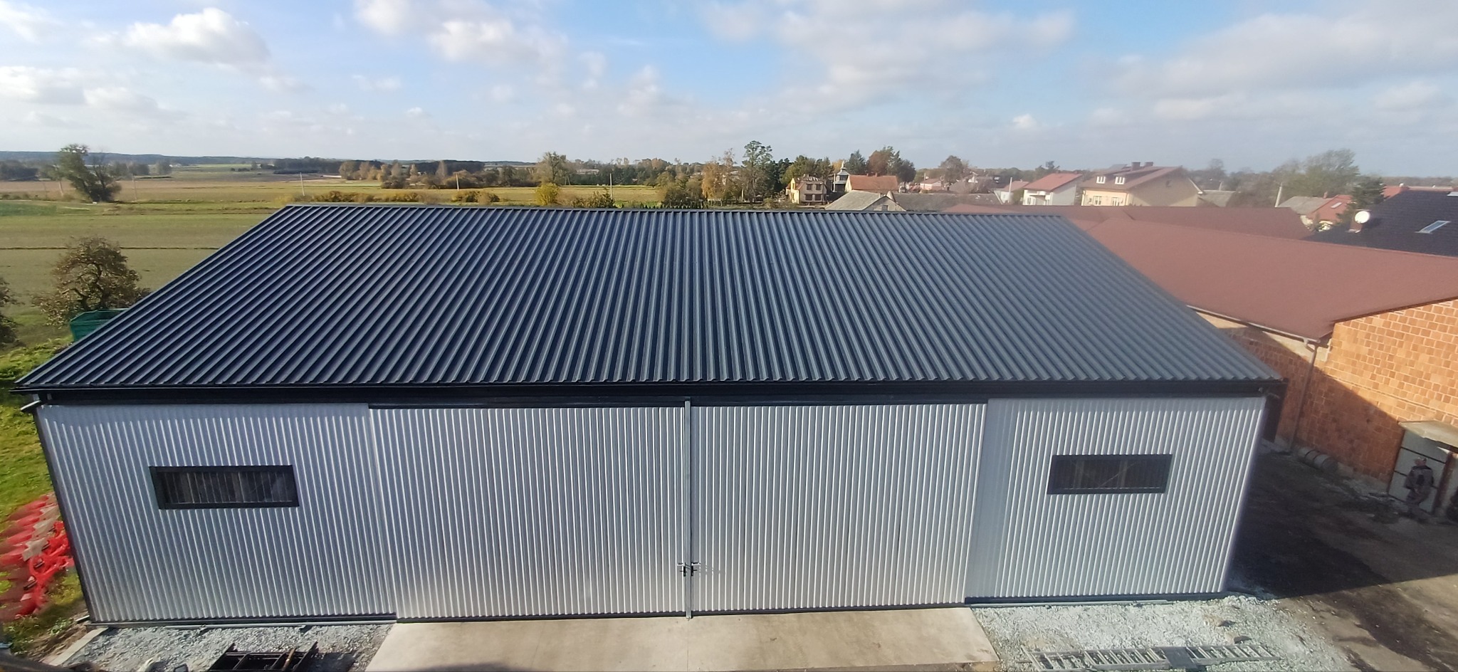 Widok z góry na nowo wybudowaną halę z blachy falistej, z ciemnym dachem i dwoma małymi oknami, na tle wiejskiego krajobrazu z zabudowaniami.