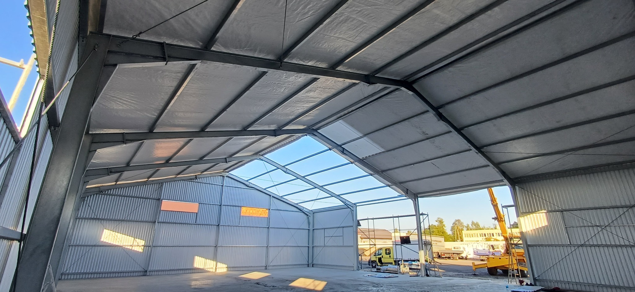 Wnętrze hali w trakcie budowy, widoczne stalowe elementy konstrukcyjne, blaszane ściany, częściowo przeszklony dach i sprzęt budowlany na zewnątrz.