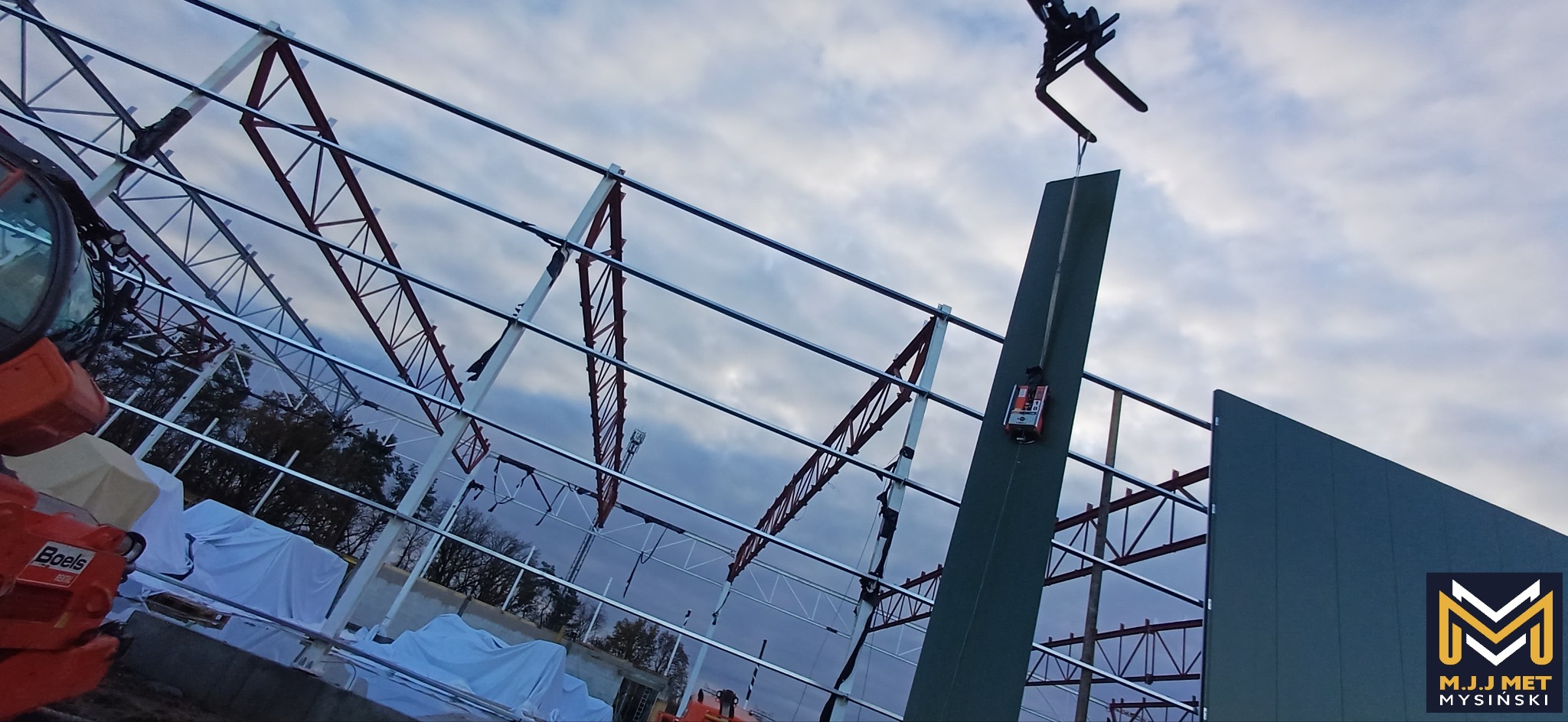 Montaż stalowej konstrukcji hali w Dąbiu z użyciem podnośnika; widoczne elementy szkieletu i obudowy, logo firmy M.J.J Met Mysiński w prawym dolnym rogu.
