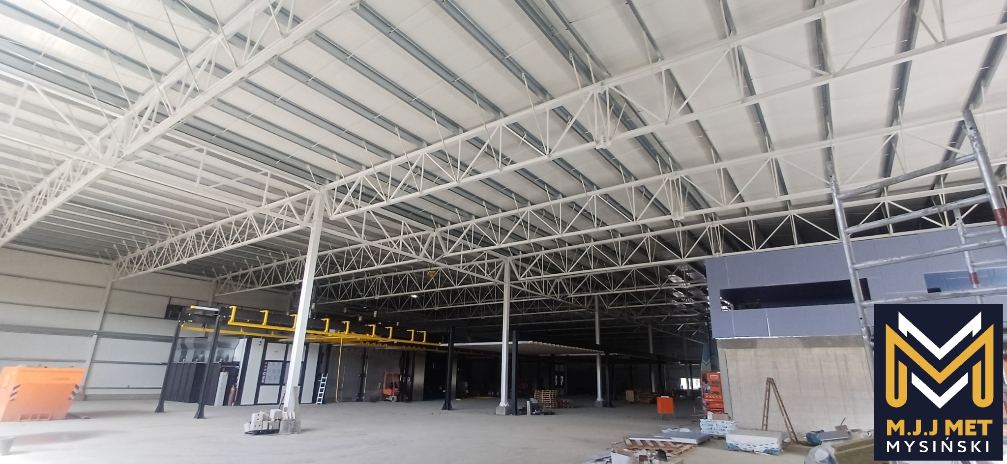Wnętrze hali produkcyjnej z widoczną konstrukcją stalową dachu, pomarańczowym kontenerem i żółtymi elementami linii produkcyjnej w tle. W prawym dolnym rogu logo firmy.