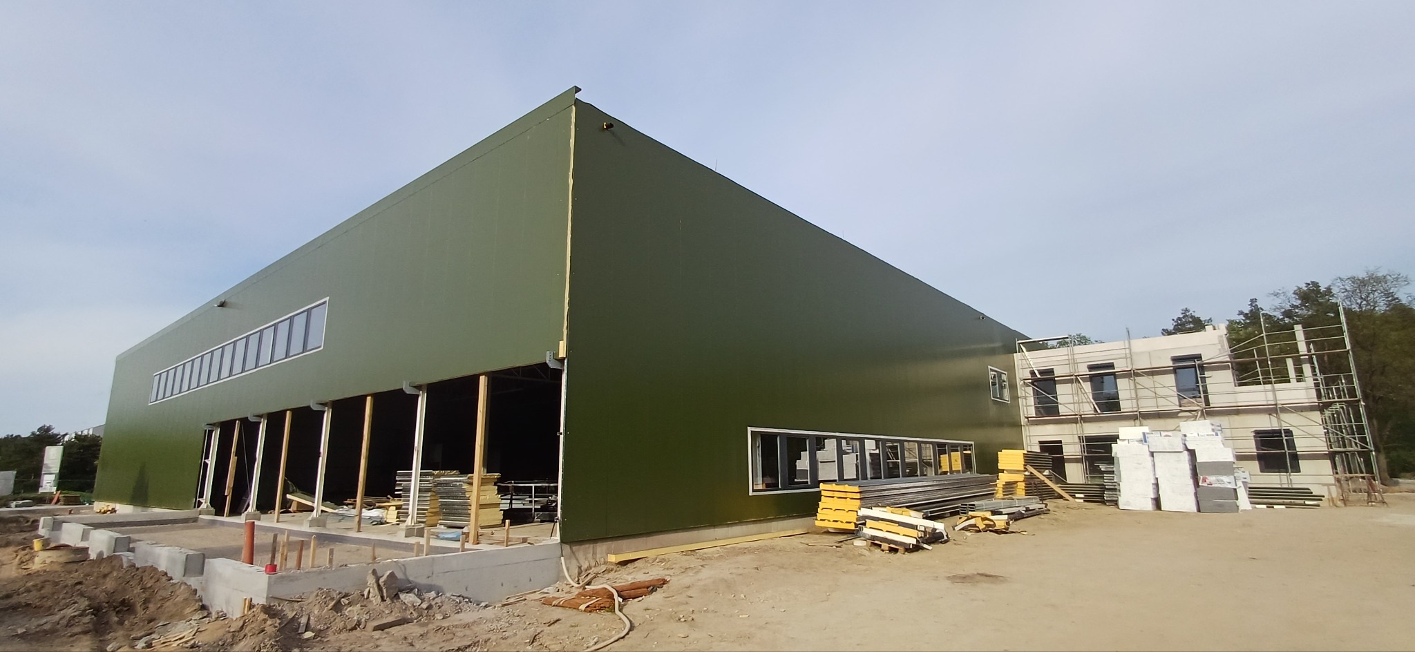 Wznoszenie hali przemysłowej z zielonymi panelami, widoczny fragment konstrukcji szkieletowej, fundamenty i materiały budowlane na placu budowy.