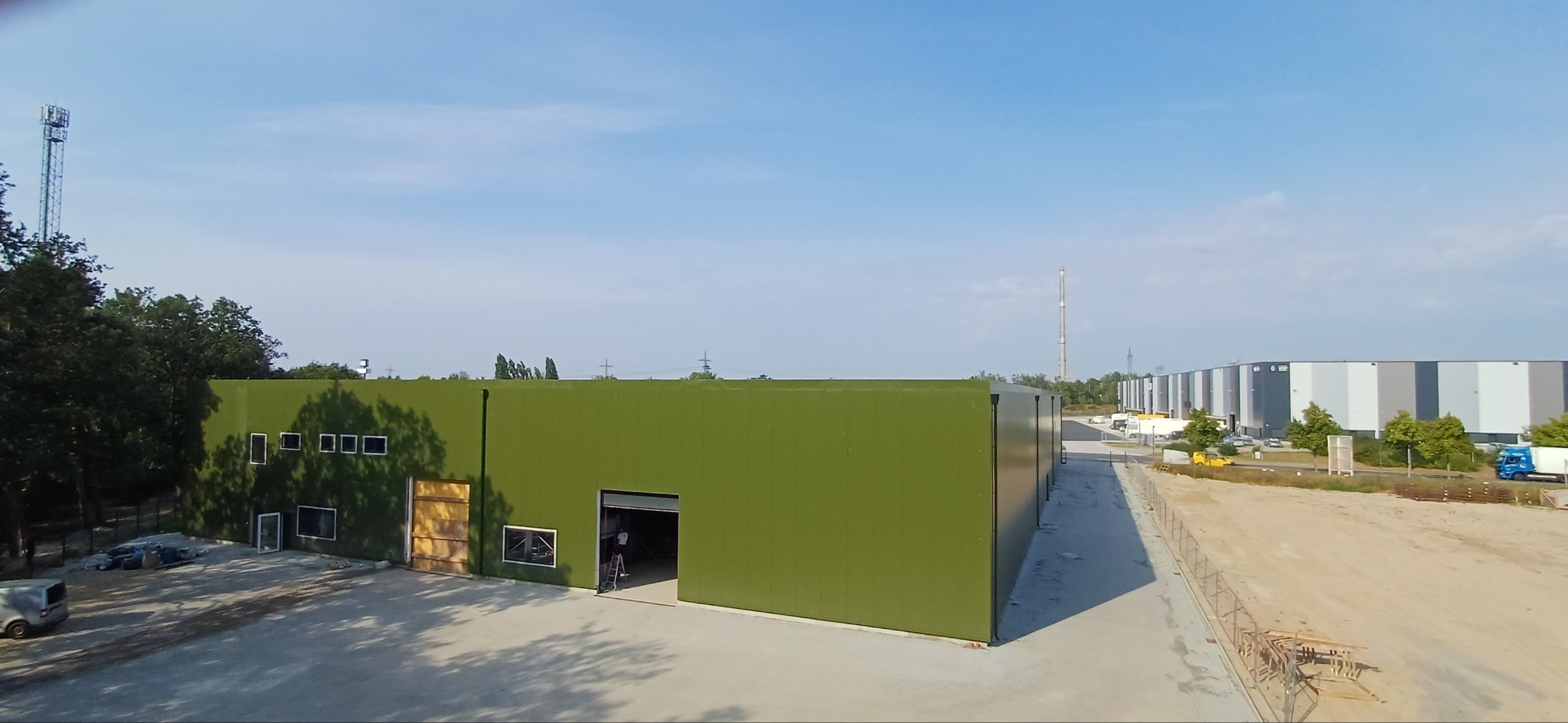 Nowo wybudowana hala w kolorze oliwkowym z otwartą bramą, widoczne rusztowanie wewnątrz, obok zaparkowany biały van, w tle inne hale i las.